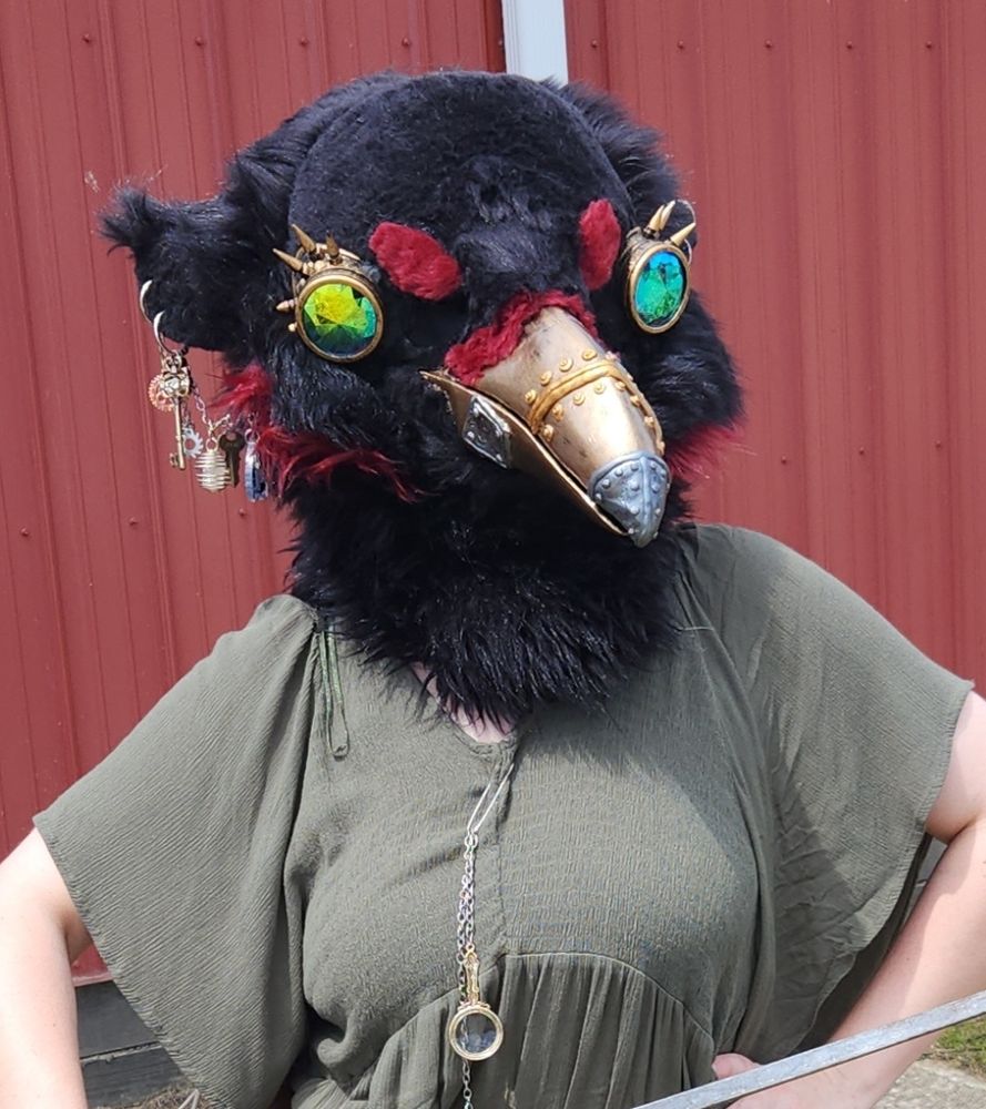 A black and red steampunk themed crow fursuit head being worn on someone with a green dress on