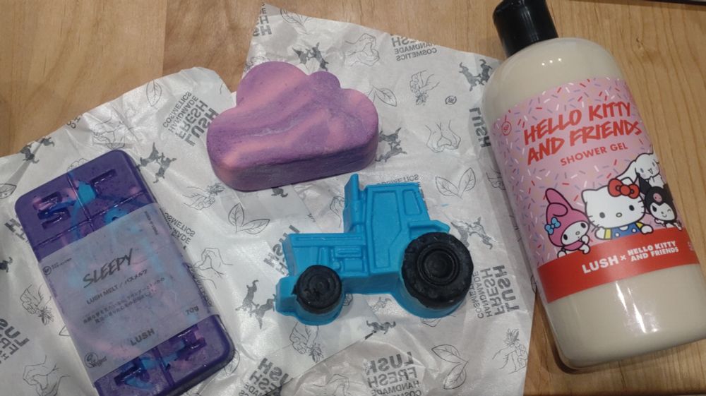 テーブルにラッシュの商品が並んでいる写真。全部で4アイテムで、左からバスメルツのスリーピー、雲の形をしたバスボム、黒いタイヤの水色のトラクターのソープ、ハローキティアンドフレンズとのコラボのシャワージェル