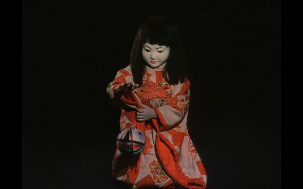 picture of a creepy Japanese doll in a red and pink kimono, playing with a ball in front of a black background from "Lullaby to Kill" (Kon Ishikawa, 1977)