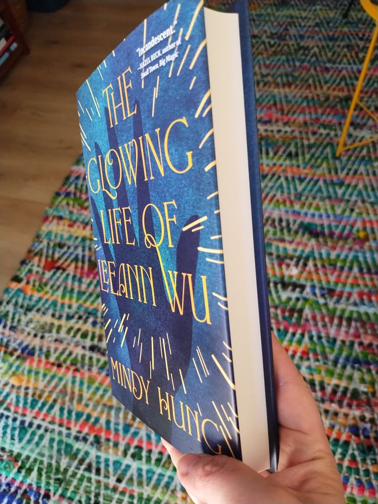 multicoloured rug in background and my hand holding a tilted hardcover copy of THE GLOWING LIFE OF LEEANN WU (so that you can see how thick it is, lol)