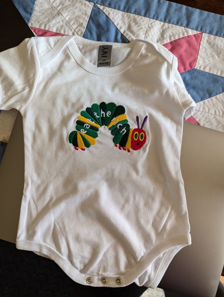A white onesie with the very hungry caterpillar and Eat The Rich written on it. 