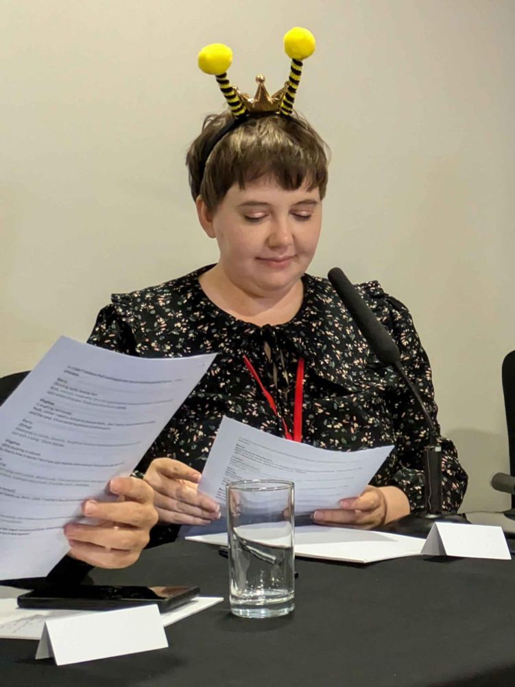 A photo of me wearing a black dress, looking down at a script, with a bee antenna on my head. Sitting behind a black table with a microphone on it.