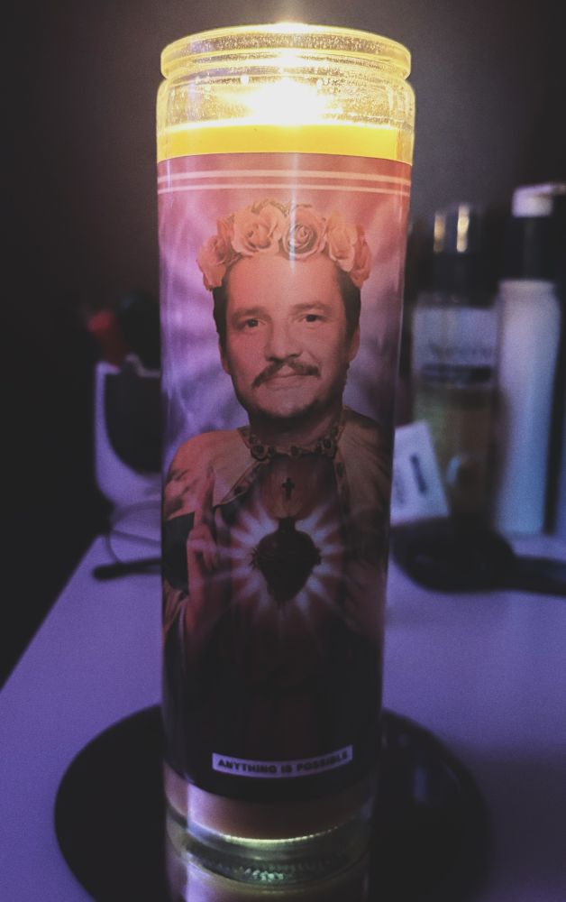A tall glass candle with yellow wax, flame visible at the top against a purple lit background. The decal on the candle vessel shows Pedro Pascal edited into a traditional prayer candle saintly figure with a flower crown and a sacred heart symbol. 