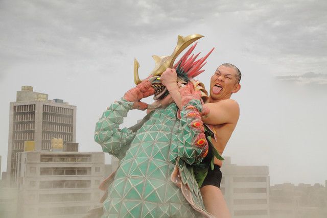 Minoru Suzuki has a big kaiju in a rear naked choke in a miniature city