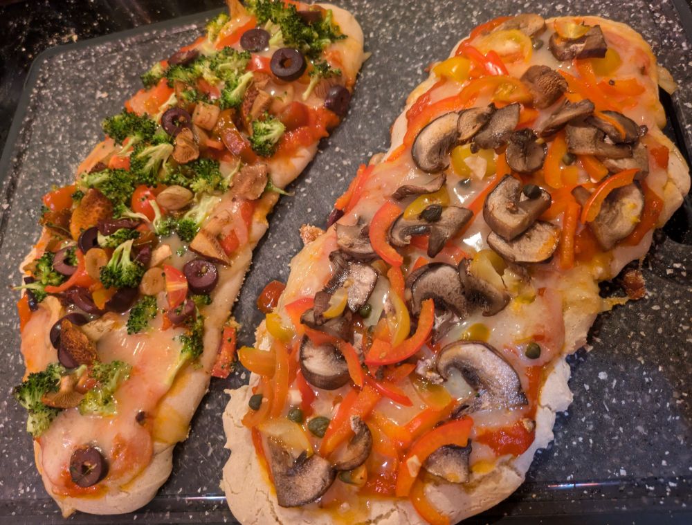 Two flatbread style pizzas on top of a grey cutting board. One pizza is primarily red with cheese and mushrooms. The other has broccoli, cheese and olives.