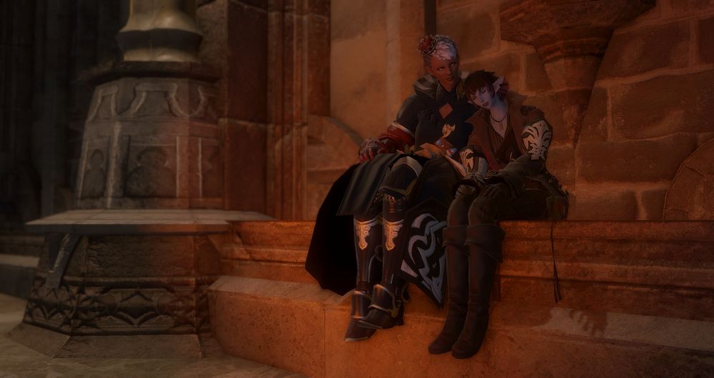 A screenshot from Final Fantasy XIV. A female Hellsguard Roegadyn and a female Duskwight Elezen sit on a stone ledge together. The Roe wears a long blue coat with gauntlets and greaves on her arms and legs, and the Elezen wears a brown coat with trousers and long boots. The Elezen's hair is trimmed and messy, and she leans her head on the Roe's shoulder, dozing off. The Roe is smiling towards the Elezen as she rests.