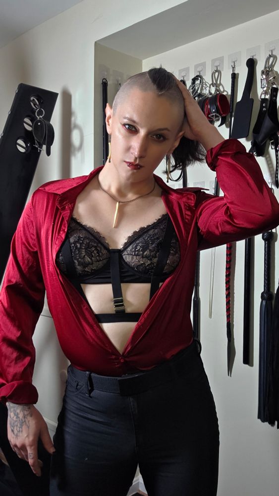 C0ffinCunt with her hair tied back. She is wearing a satin red button-down, a harness bra with lace, a baculum bone around her neck, raccoon finger bones in her ears, res lip stain, and black & red eyeshadow.