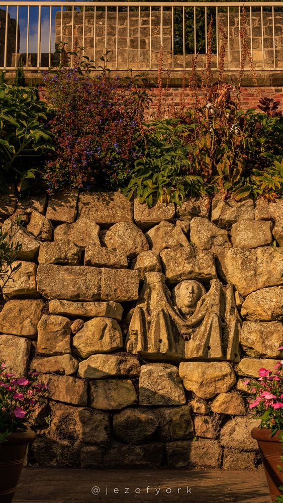 A carved stone face set into a stone wall topped with plants sticks out its tongue at the viewer. A second castellated wall sits above and behind.