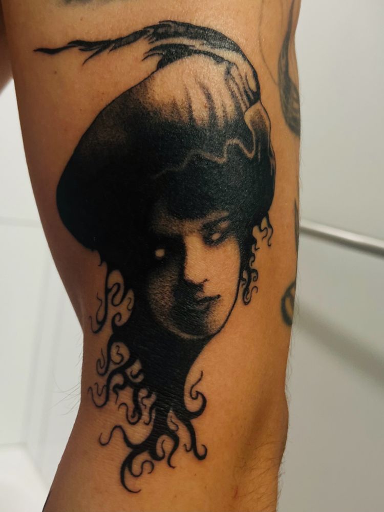Black and gray tattoo of a Victorian portrait of a woman with a feather hat, dark hair coming down and forming tribal styled tendrils. Done by Cris Crude at State of Love Tattoo in Richmond, VA