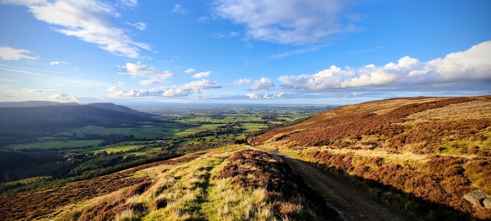 A wide vista looking out from on top of a tall hill. More hills sit at the sides, a flat valley stretches into the distance where a faraway range of mountains just can be seen. In the foreground the remaining earthworks of a Victorian railway track can be seen running down the steep hill.