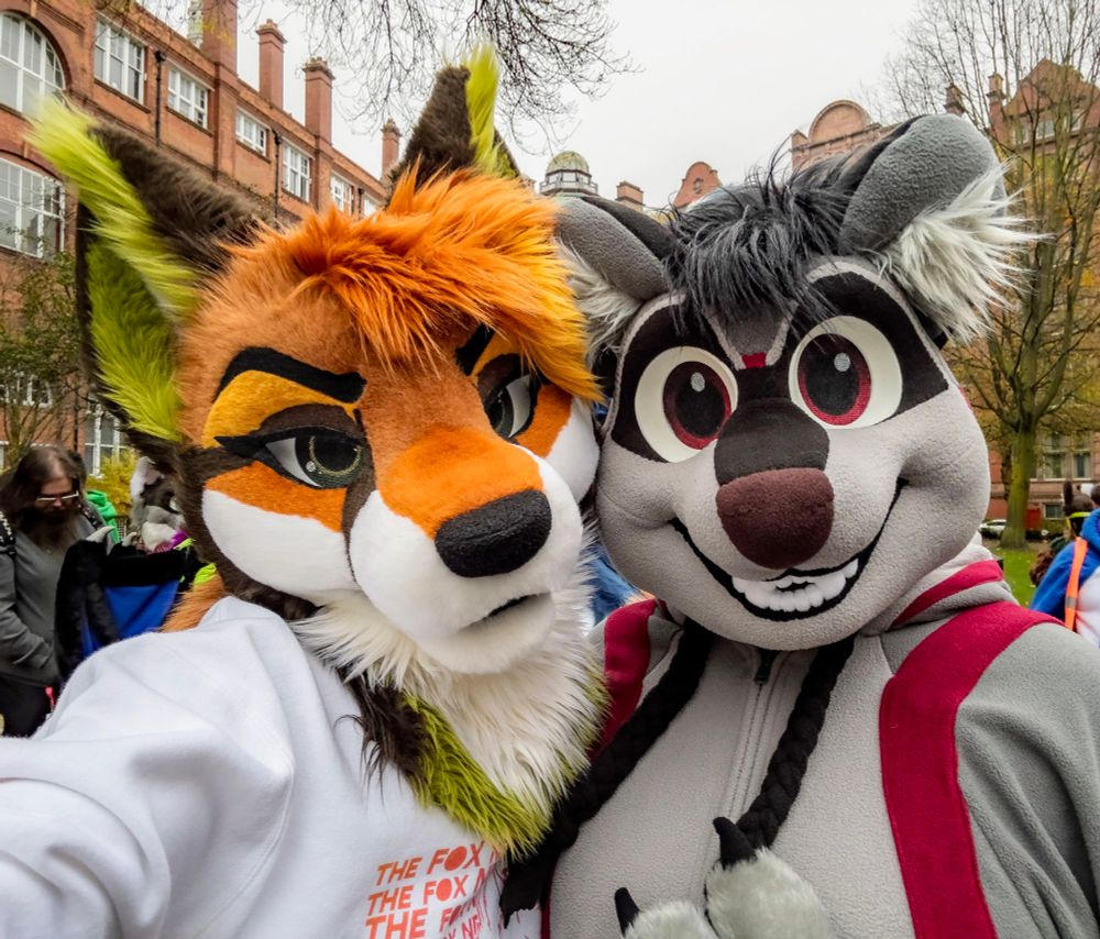 Two fursuiters taking a selfie, one is a fox in a comfy jumper and the other a raccoon in a fleecy Kigu