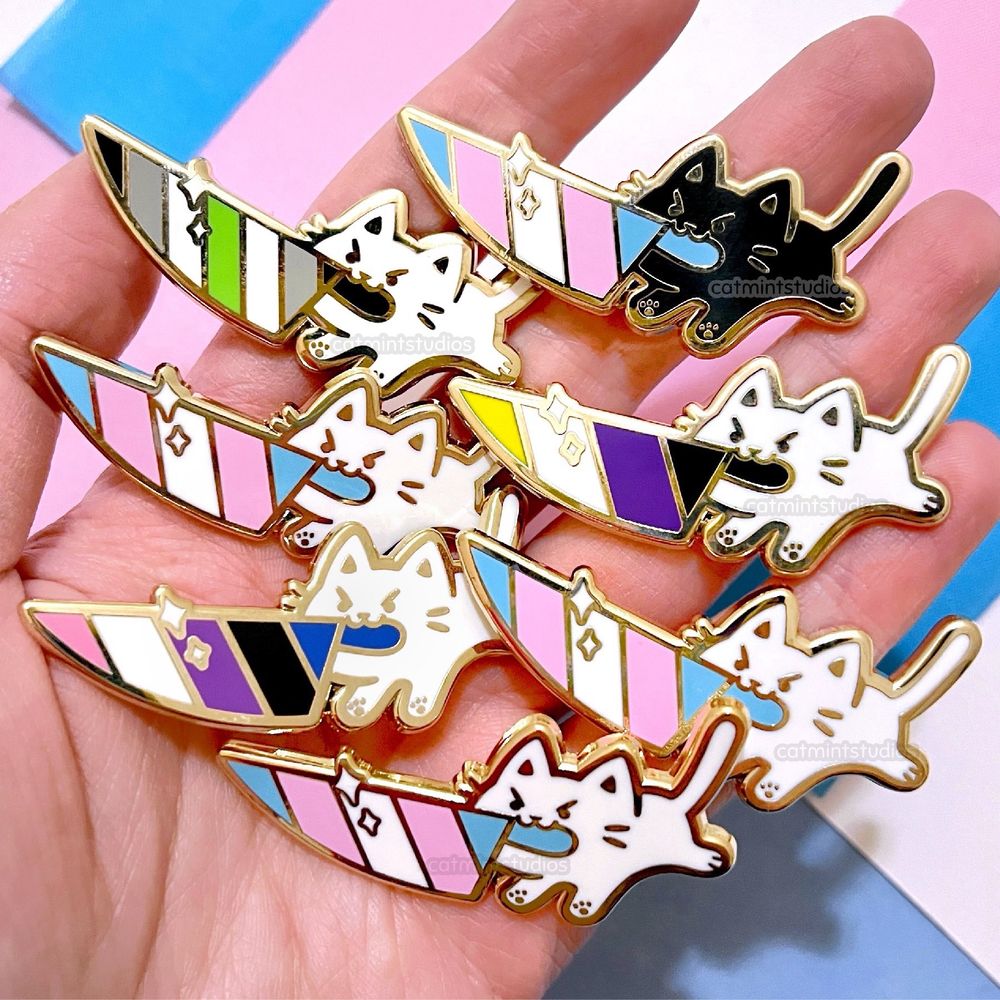 A hand holds enamel pins over a trans pride flag background. The pins are all small cats (mostly white cats with one black cat) holding knives in their mouths that have pride flag designs inside. The flags are transgender, genderfluid, nonbinary, and agender.