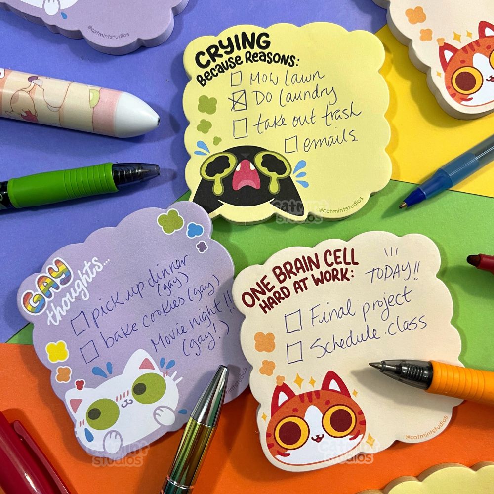 A series of 3 sticky note pads sit on a purple, green, yellow and orange background with various pens scattered around. The first design is a light purple pad with a white cat face at the bottom. The cat face has green eyes and has their paws up looking nervous with a little blush and sweat drops. They have a thought bubble above their head that says “Gay thoughts…” 
The second design is a light yellow-green pad with a black cat face crying at the bottom. On the top of the pad it says “crying because reasons…”
The last design is a beige pad with a goofy looking orange tabby cat face at the bottom with his tongue out. On top of the pad it says “one brain cell hard at work…”