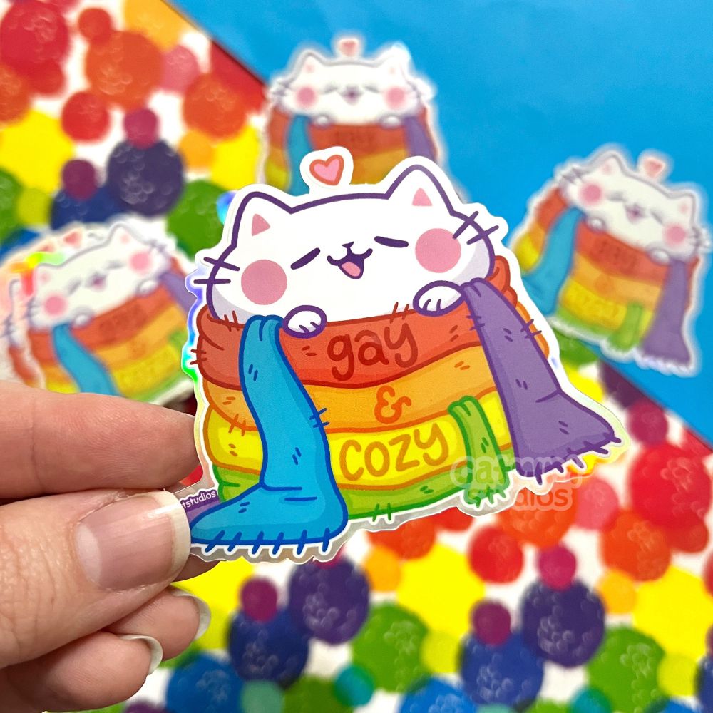 A hand holds a sticker above a rainbow polka dot and blue background. The same sticker is scattered in the background. The sticker design is of a cute chubby white cat wrapped in a rainbow scarf. The words “gay & cozy” are written on the scarf. 