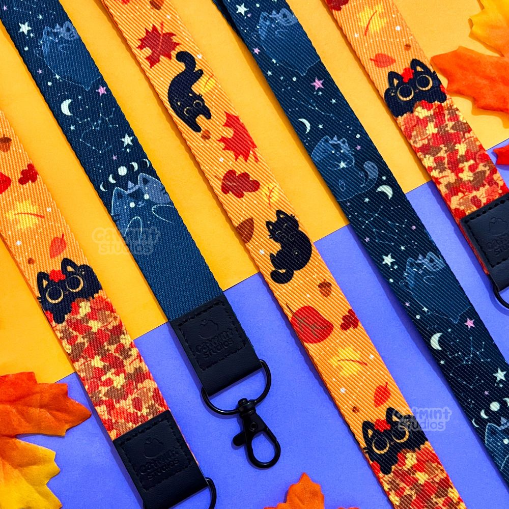 2 lanyards lay on a yellow and purple background with autumn leaves scattered around them. The first lanyard is primarily orange with red, brown, and yellow leaves and features different poses of a cute black cat. The black cat is playing in a pile of leaves at the bottom of the lanyard. The second lanyard design is primarily dark blue/black with a black and gray witch kitty. There are stars and constellations in the background. The kitty at the bottom of the lanyard has the moon phases above its head. Both lanyards have a black leather section at the end with the clasp that has an embossed Catmint Studios logo. 