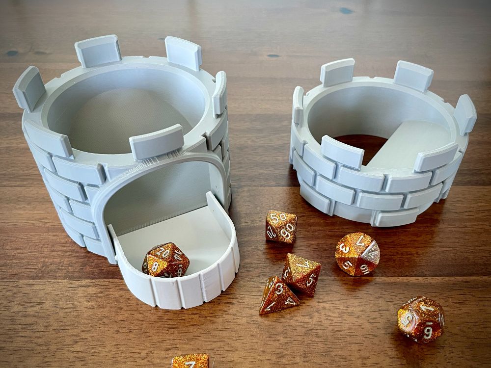 Two parts of a 3d printed dice tower, on the left is a short medieval-style stone tower with lowered drawbridge forming a tray, with a D20 inside. Crenelations along the top fit the bottom of the piece on the right, an insert that adds another level and ramp for die to bounce off. Identical crenelations are on the top of this piece.