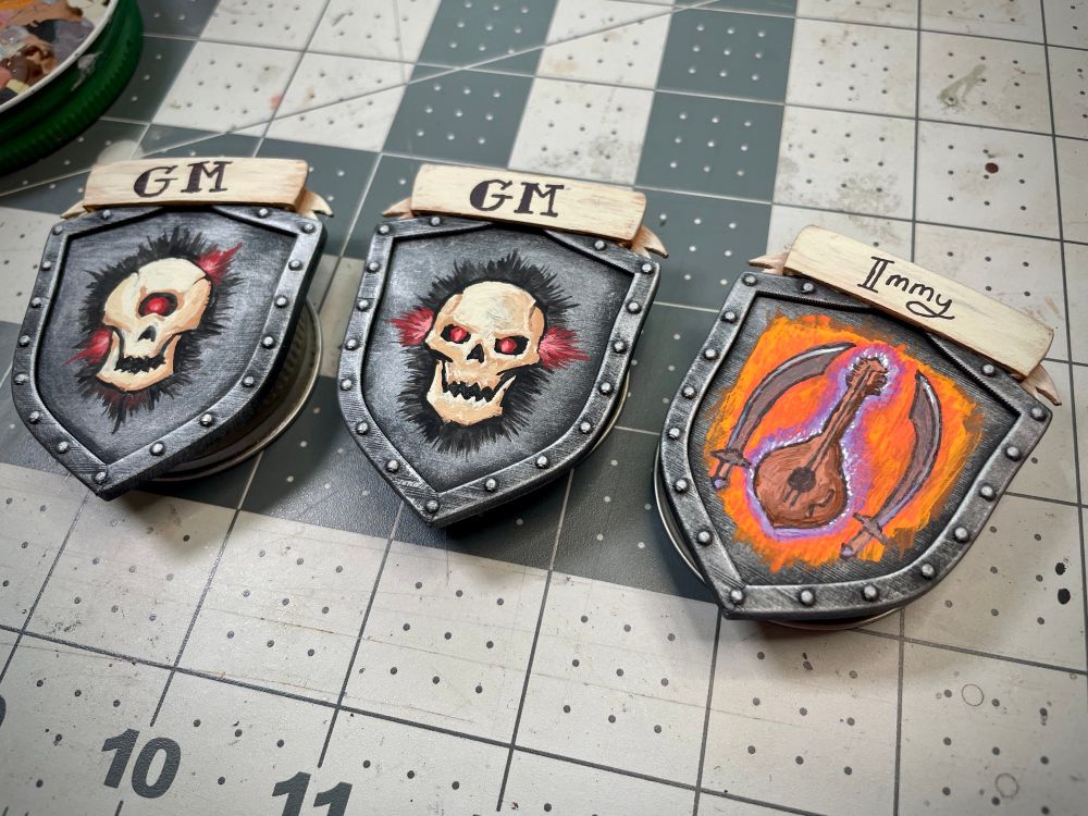 Three miniature painter tokens of shields with banners across the top. The first had a cyclops skull with glowing red eye painted on the shield, with GM written on the banner. The second also has GM on the banner, but painted on the shield is a human-like skull with glowing red eyes. The last token seems to be for a player, with lighter text on the banner displaying the name ‘Immy’. The emblem on this shield is surrounded by fiery orange and depicts two scimitars flanking a lute wreathed in purple flame. 