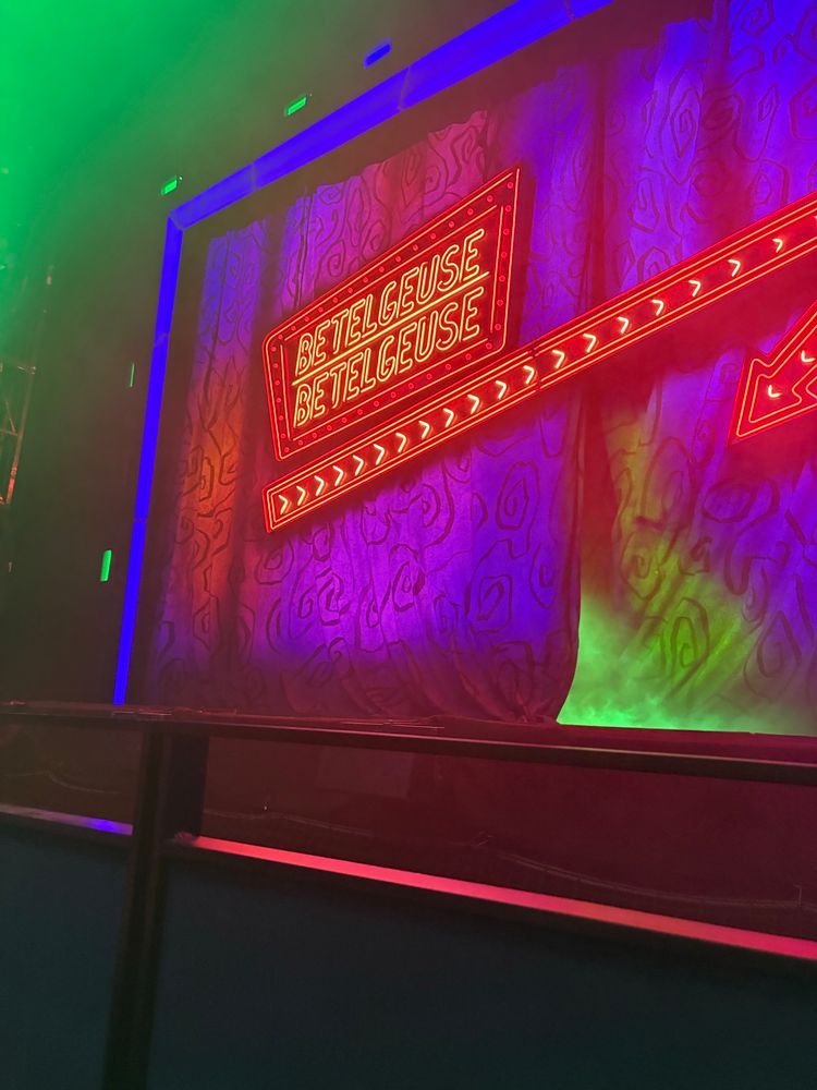 a view of a stage from the front row. It’s foggy, lit in multiple super vibrant colours and has an angular neon sign that reads “Betelgeuse Betelgeuse” with an arrow.