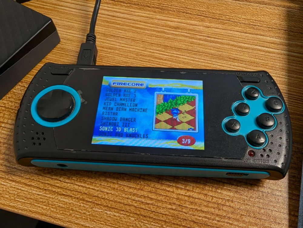 A handheld AtGames system with a black and blue color scheme, there are visible signs of wear from years of heavy use, the system is plugged into a computer to charge, and the screen shows a list of games with Sonic 3D Blast highlighted