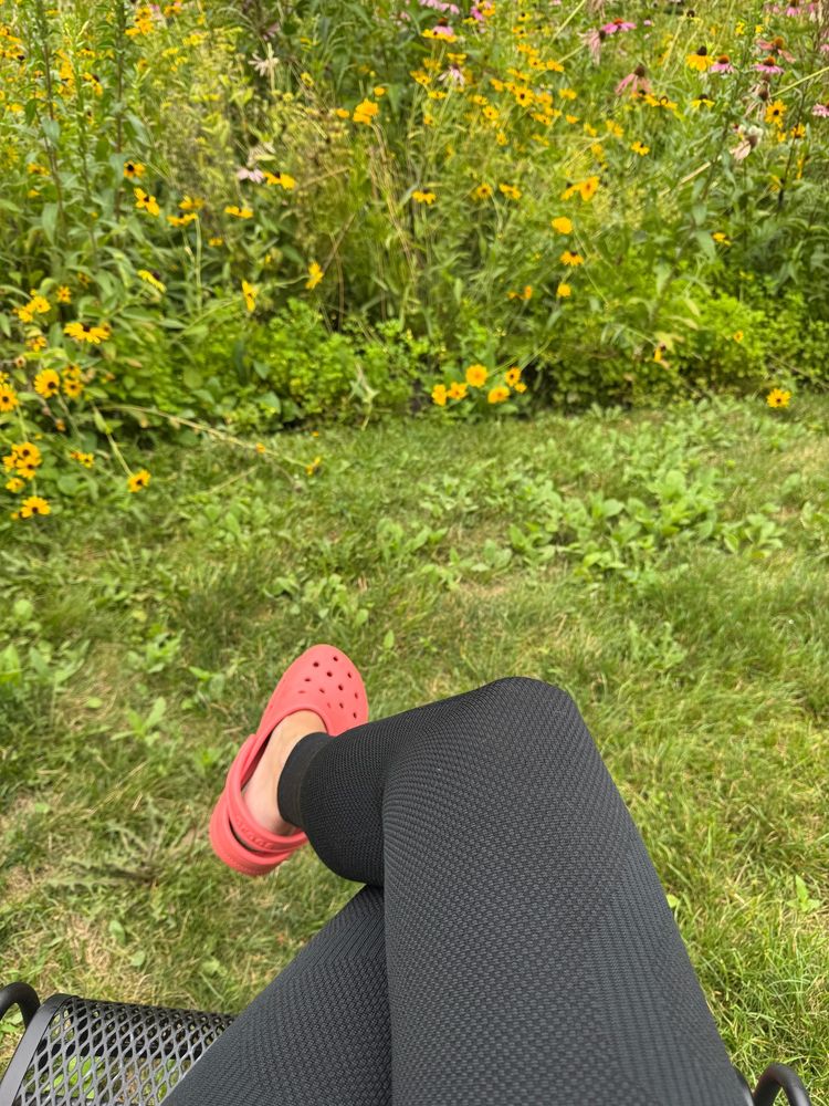 Selfie of my legs, seated in a chair next to the garden. I’m wearing black compression leg sleeves and coral crocs