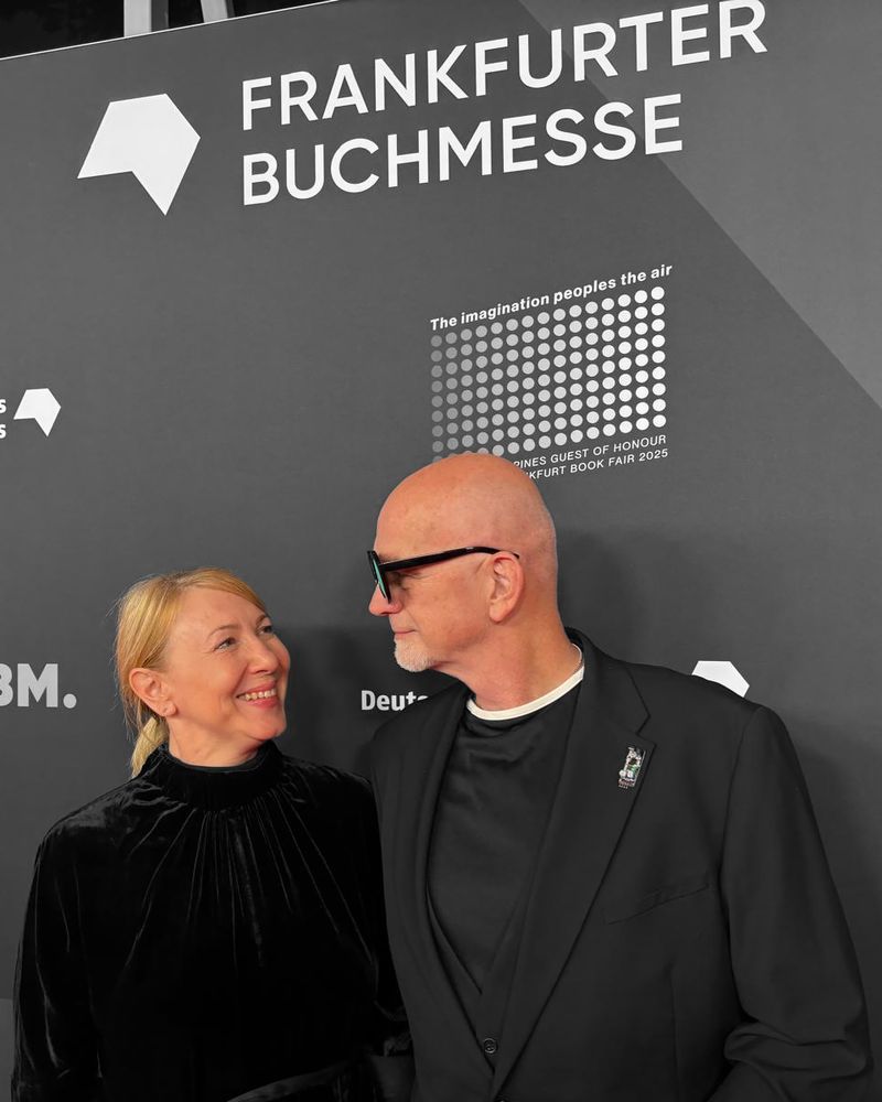 A woman and a man stand close together in front of the Frankfurter Buchmesse backdrop. They are smiling at each other. The woman wears a black velvet dress, her blonde hair tied back. The man, bald with a short white beard, wears black-framed glasses and a black suit with a small lapel pin. The gray background features the white text and dots of the Frankfurter Buchmesse 2025 design.