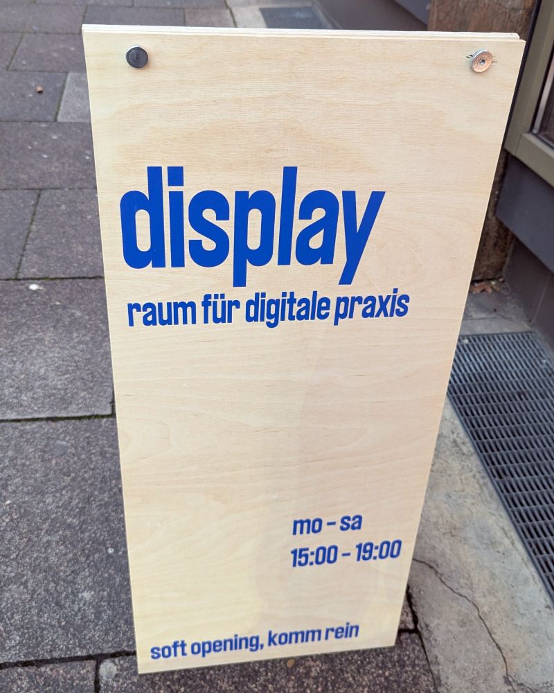 A freestanding wooden sidewalk sign stands on a city pavement. It has bold blue text that reads: "display raum für digitale praxis." Below it, smaller text lists opening hours: "mo – sa 15:00 – 19:00." At the bottom, it says "soft opening, komm rein."