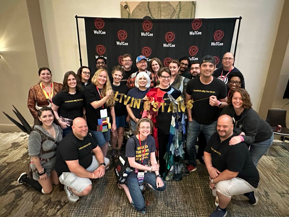A group of Michael Livingston fans at WoT Con gather together smiling and holding a gold Living Stans banner.