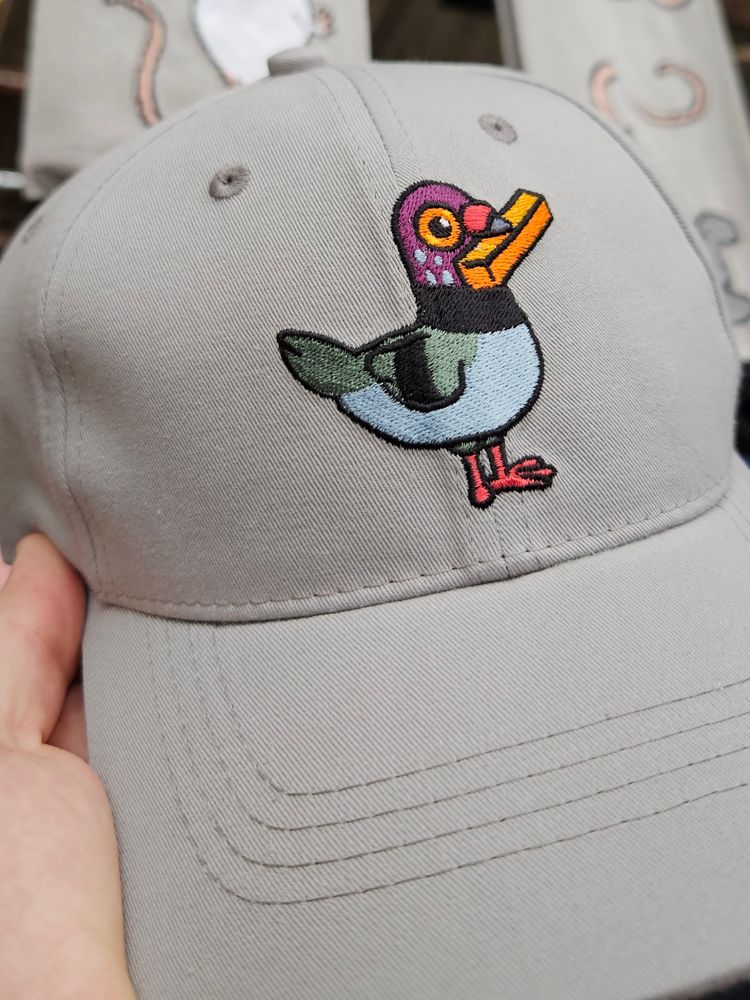 Un pigeon brodé sur une casquette grise. Il tient une frite dans son bec.