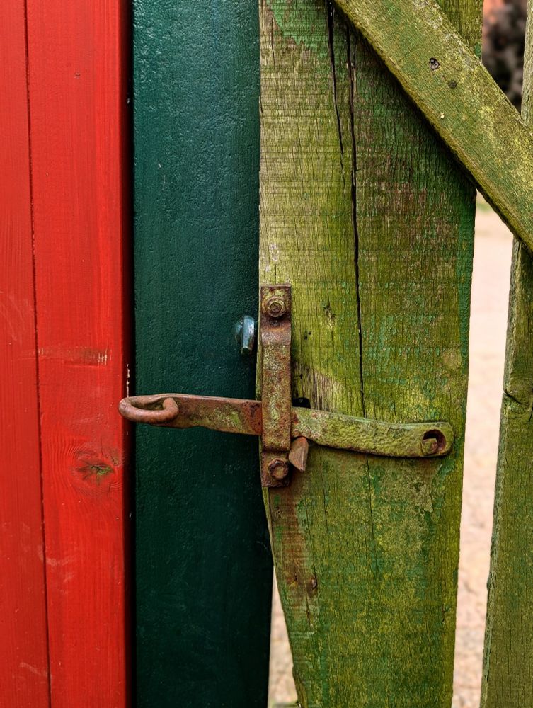 Das Foto zeigt einen alten verrosteten Türhebel an einem bemosten Tor. Die Pfosten links daneben sind dunkelgrün und rot