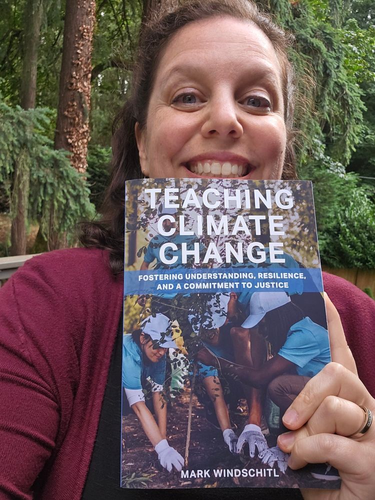 Woman with brown hair and maroon sweater standing in front of evergreen trees holding a book just below her smiling face. The cover of the book shows people planting trees with a title Teaching Climate Change Fostering Understanding, Resilience, and a Committment to Justice by Mark Windschitl