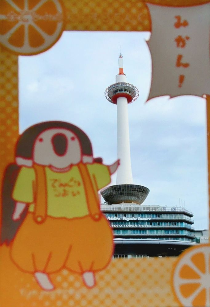 てんぐの子どもとみかんの描かれたアクリルキーホルダーの写真。中央が透明で、ニデック京都タワーが透けて見える。てんぐの子どもは中央に指をさし、「みかん！」と叫んでいる。