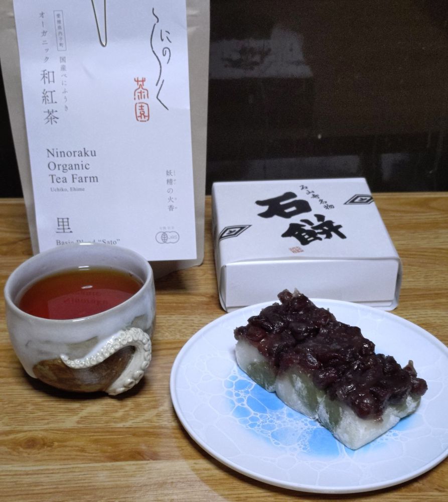 お茶とお餅の写真。手前右にあんころ餅ののったお皿、その奥に「石餅」と書かれた白い紙箱、手前左にお茶の入ったフリーカップ、その奥にお茶の白い袋パッケージが置いてある。フリーカップは白いイカの足がカップの側面に絡みついているようなデザイン。