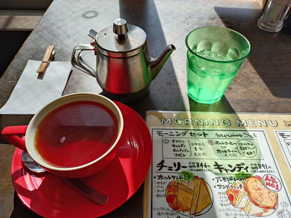 紅茶の入ったティーカップと喫茶メニューのラミネートの写真。メニューには「モーニングメニュー」として「モーニングセット　チェリー　キャンディ」の手書き文字と、メニューのイラストが描かれている。チェリーはホットサンド、キャンディはアーモンドトーストとサンドイッチ。