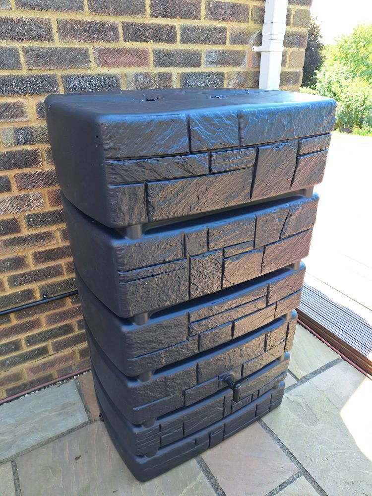 An anthracite black patio water tank with brick effect moulding on a sandstone patio, which leads to a large decking area