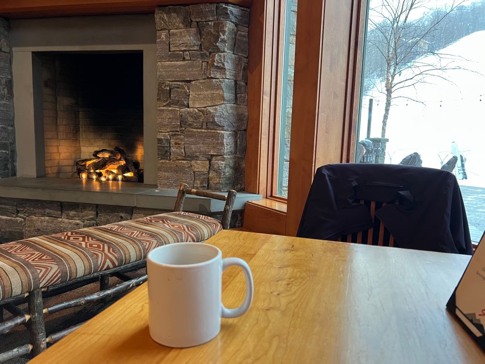 Giant white coffee mug at a wood table, overlooking a large fire place and the ski slopes. 