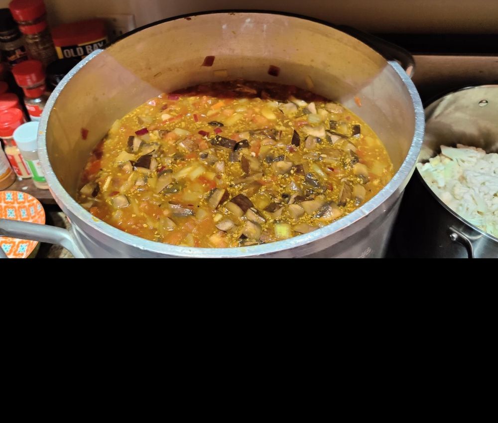 a very large pot of curry soup sits on a stovetop, with many vegetables and split peas. next to the pot of soup is  another, smaller pot filled with cauliflower, not yet cooked. on the other side is a counter with a full spice rack.