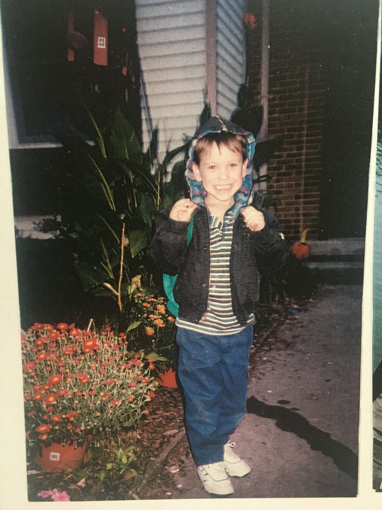 Picture of lil prof standing on his sidewalk about to head to preschool wearing a gripped shirt and jeans with a black rain jacket on and a backpack 