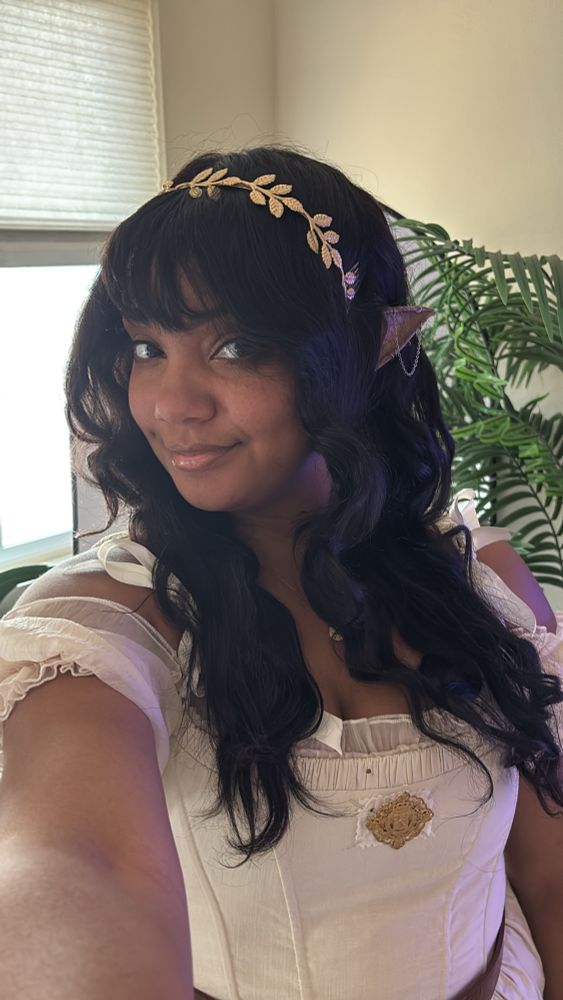 A selfie of TiffsChateau wearing a cream corset dress, over a chiffon top. She has long curly hair with bangs and elf ears. Her head is turned to the side while looking at the camera. Elf ears are peaking out.