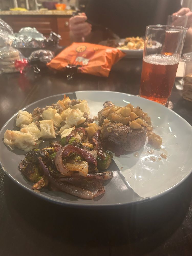 A photo of a gray and white ceramic dinner plate with Thanksgiving dinner on it: stuffing, roasted red onions and Brussel sprouts, and a pork chop topped with an apple and onion sauce. There is a glass of bubbly pink drink next to the plate.