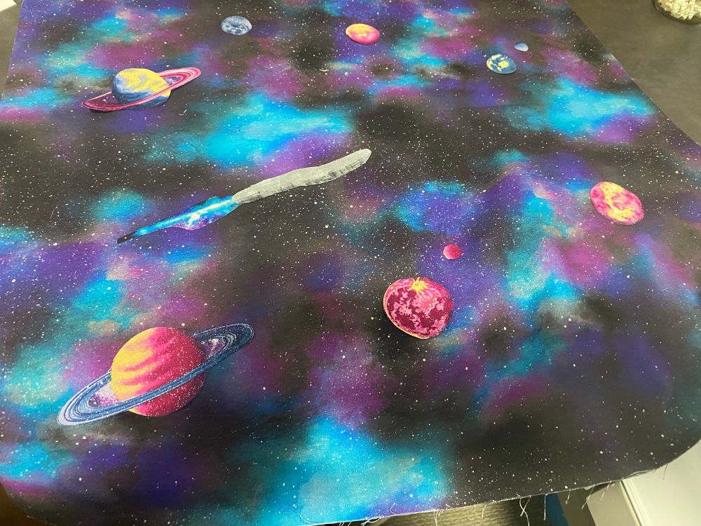 Nebula background fabric with stylized planets and space objects fused to it.  Oumuamua is created from gray fabric with permanent marker ink added to make it look like a rougher surface.  I added a comet tail to represent the newly discovered visitor 3I/Atlas comet.  Earth is depicted in top right corner along with our moon. 