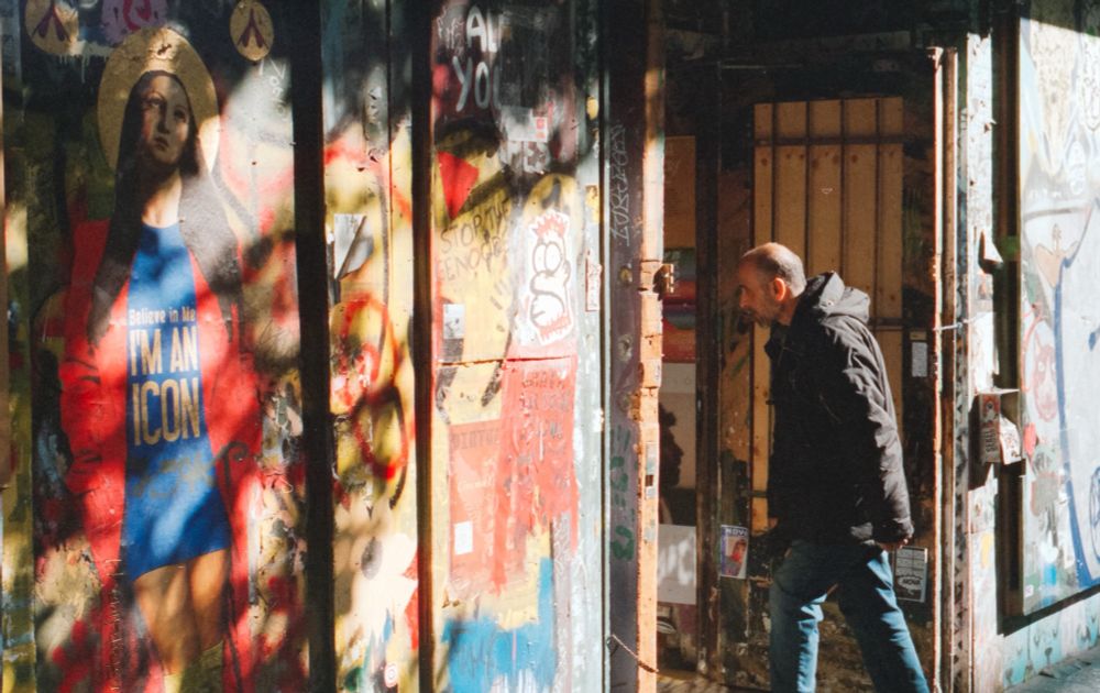 A man walking into a store, illuminated from behind by the sun, thats also basking the store front highlighting various paintings, prints and tags on the wall.