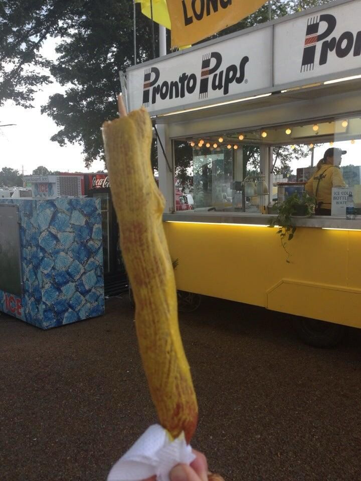 Someone holds a Memphis delicacy known as a Pronto Pup (a superior version of a corndog) with a layer of yellow mustard in front of a yellow Pronto Pup stand.