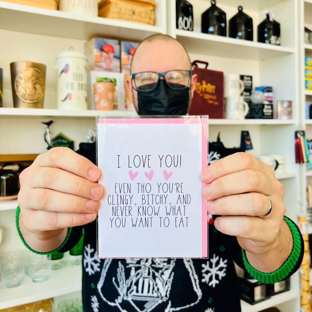 Meg’s spouse holds a greeting card that says “I love you even though you’re clingy, bitchy, and never know what you want to eat.”