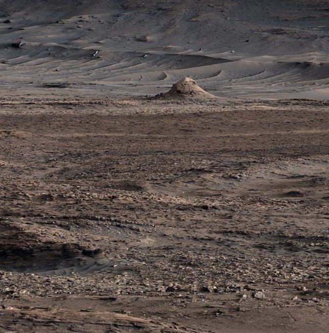 A photo from NASA's Curiosity rover showing a desert landscape with rough, rocky terrain in the foreground, dunes at the base of a hill in the background, along with a relatively small, layered hillock of sandstone. 