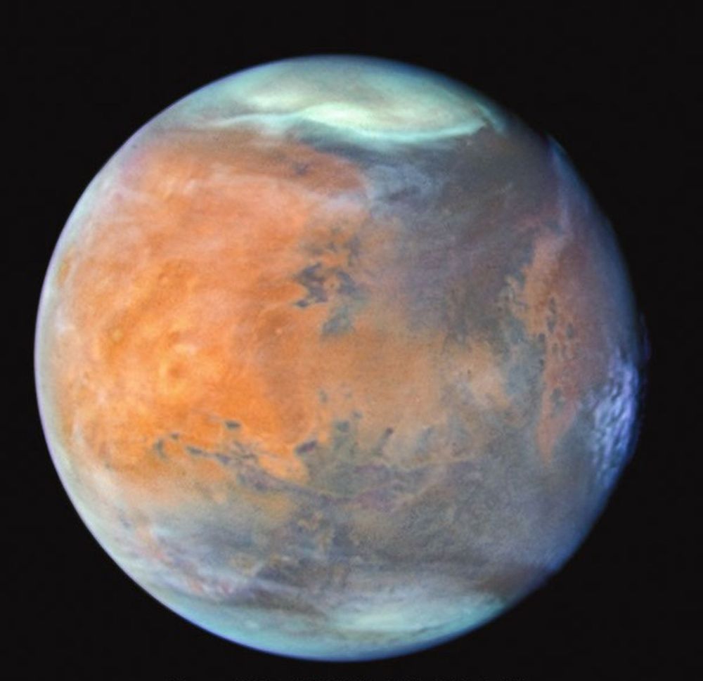 A full-disc view of the planet Mars observed by the Hubble Space Telescope against a black background of space. The planet's surface has a red-orange tint. White clouds obscure much of the polar regions. Other clouds and haze drift through the atmosphere. Dark patches of exposed rock, craters, plains, and volcanic mountains can be seen.