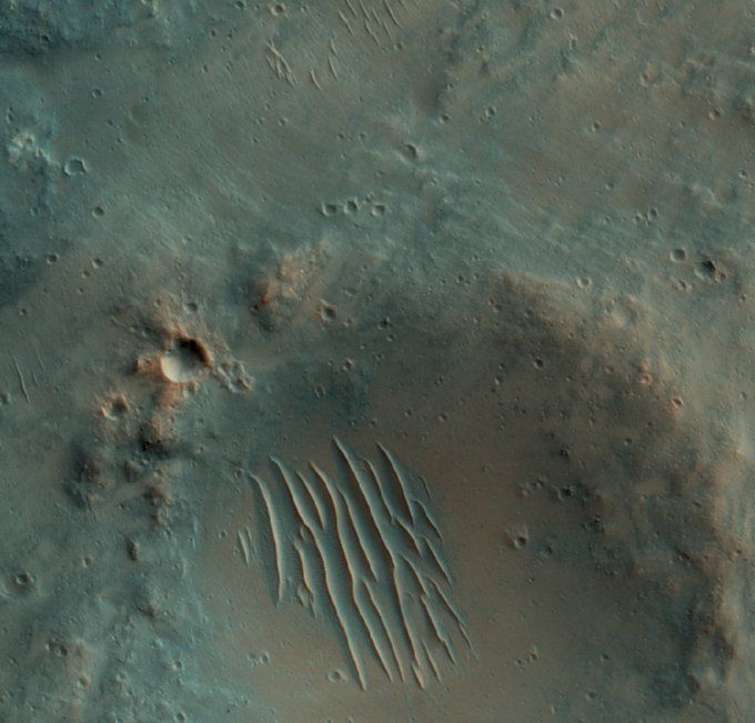 A photo from NASA's Mars Reconnaissance Orbiter with a shallow crater seen from above and parallel lines of small dunes within it. Other, smaller craters dot the landscape as well. 
