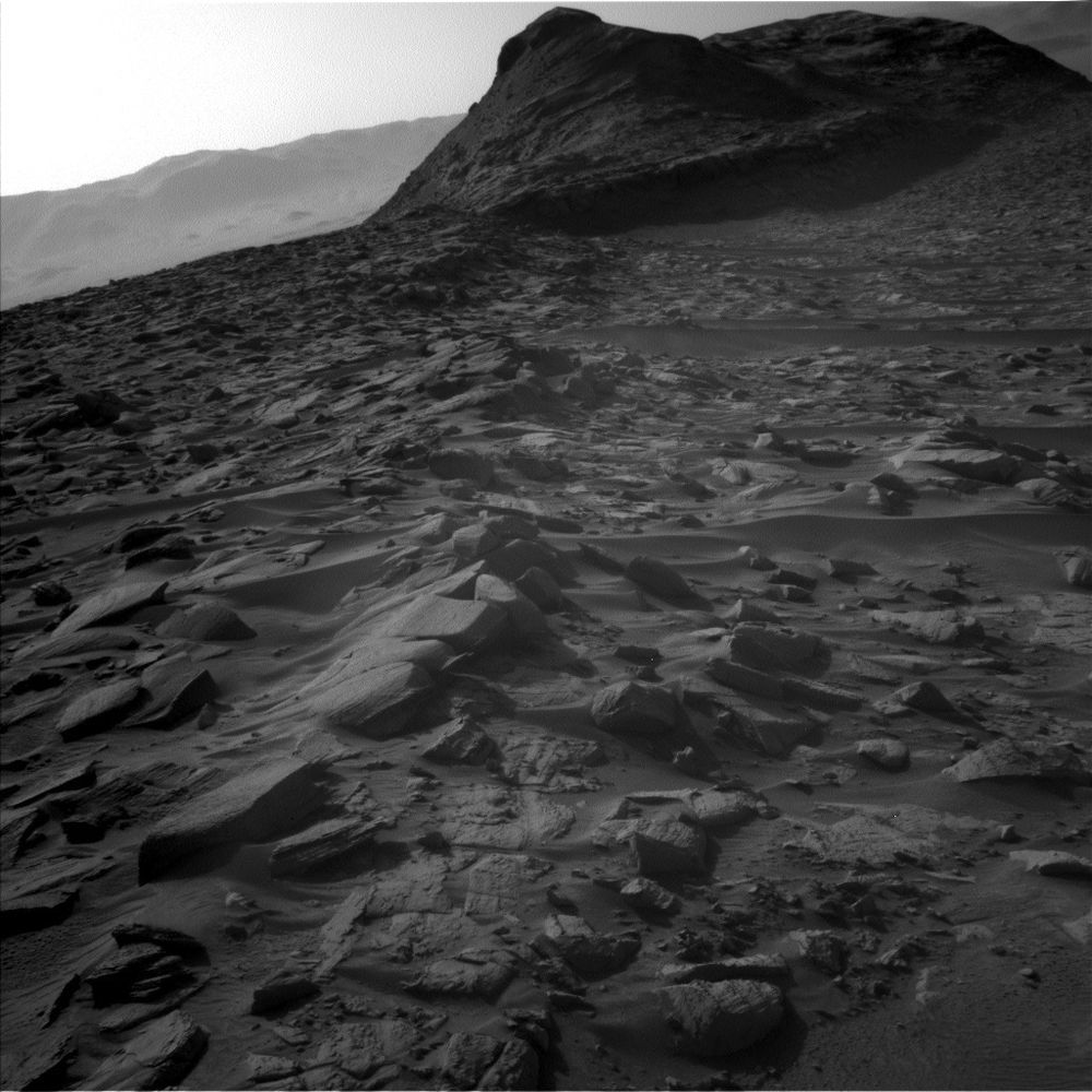Black-and-white photo from the Curiosity rover of a rocky desert terrain, shadowy in the low sunlight. Scattered rocks and ripples of dust in the foreground. In the middle distance rises a layered hill of rock. The crater rim appears as a mountain range on the distant horizon.
