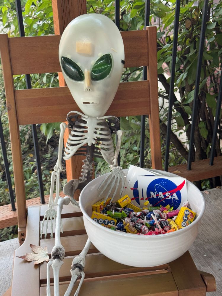 A Halloween display skeleton wearing an alien mask, next to a candy bowl with candy and a stack of NASA stickers 