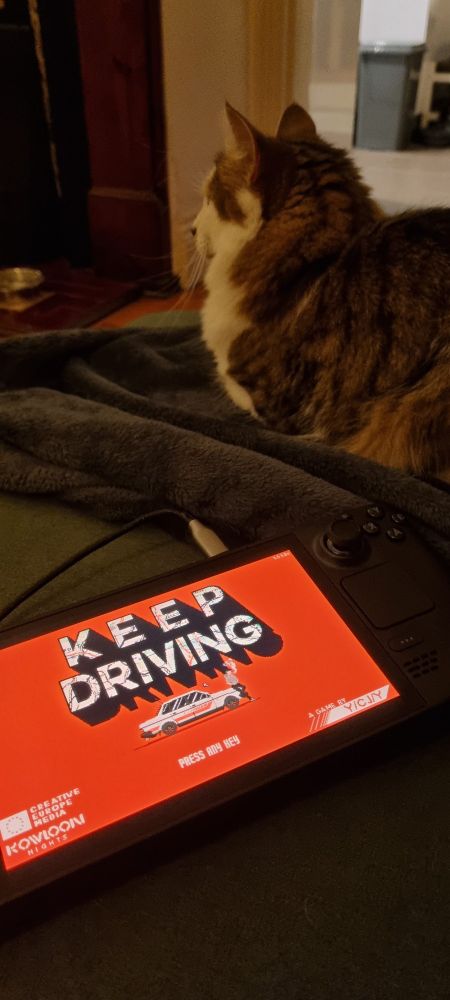 A cat sits next to a steamdeck in the foreground shows the start screen for Keep Driving.