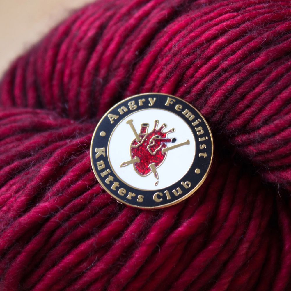 An enamel pin of an anatomical heart with knitting needles poked through it that says angry feminist knitters club